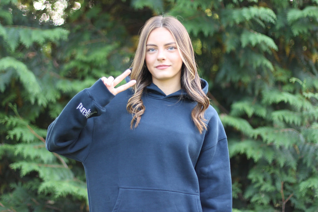 Person wearing a navy hoodie with 'Purelight' branding, making a peace sign in front of green trees.