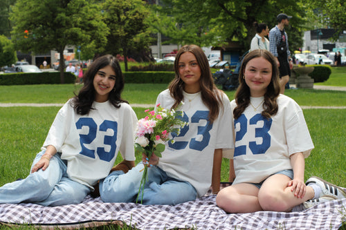 Three women sitting on a blanket outdoors, wearing matching shirts with the number 23.