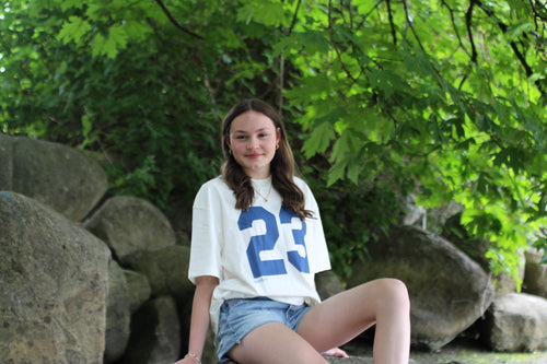 Young woman wearing a white t-shirt with the number 23 sitting among rocks and greenery.