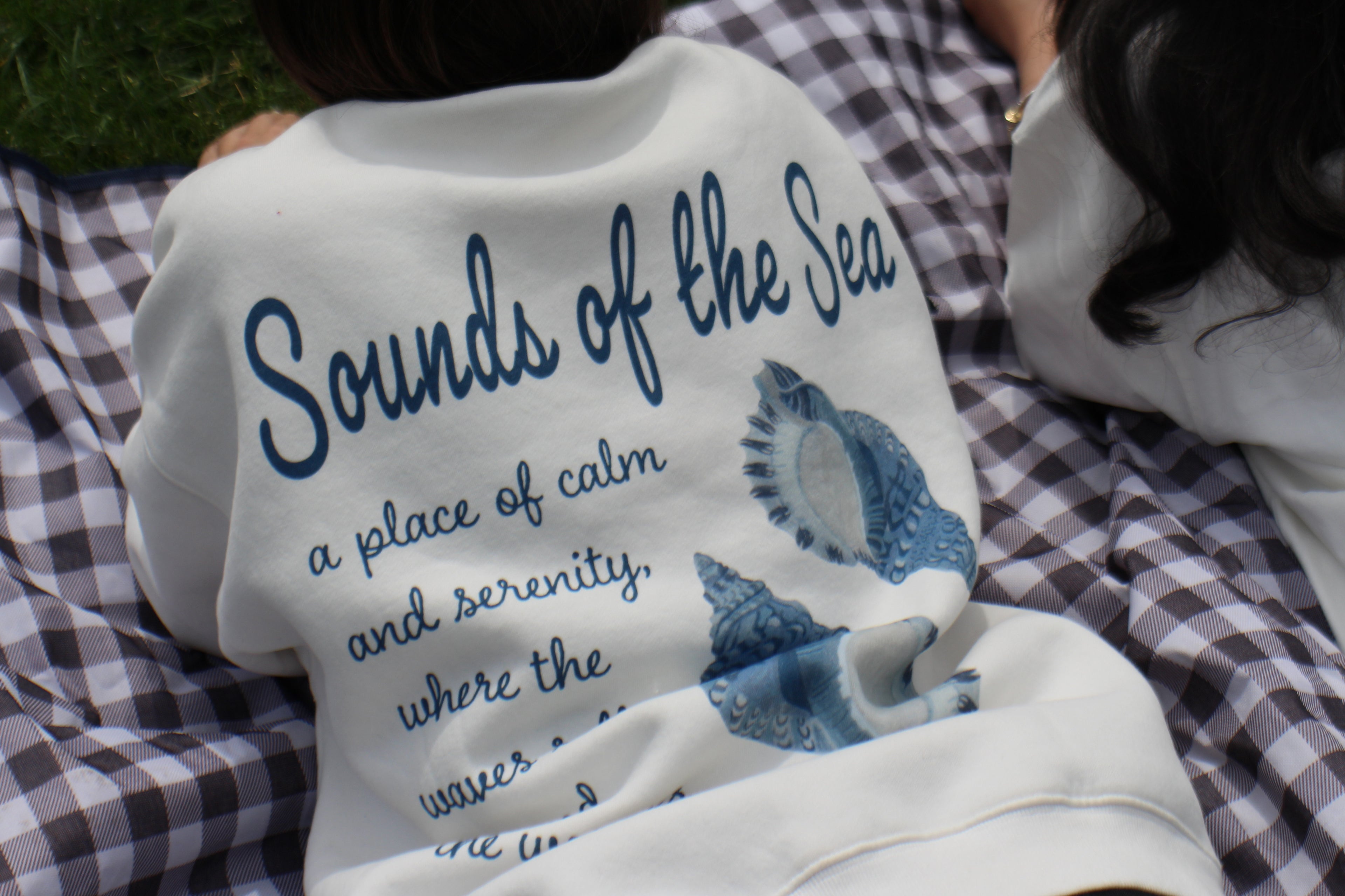 Person wearing a shirt with 'Sounds of the Sea' text and design, sitting on a checkered blanket.