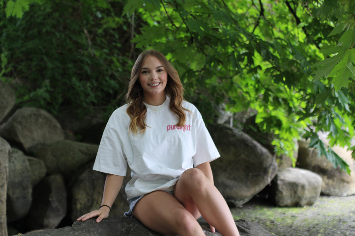 Person wearing a white t-shirt with 'puregift' logo sitting outdoors among rocks and greenery